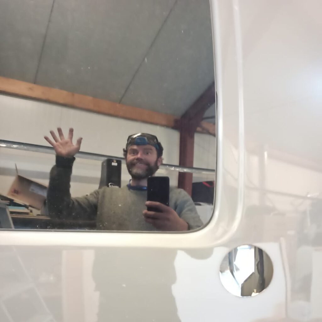 Selfie van Emile van Tiny House Campers met een hoofdlampje op, zwaaiend naar zichzelf (dus naar ons) in de spiegeling van een van de ramen van de bus