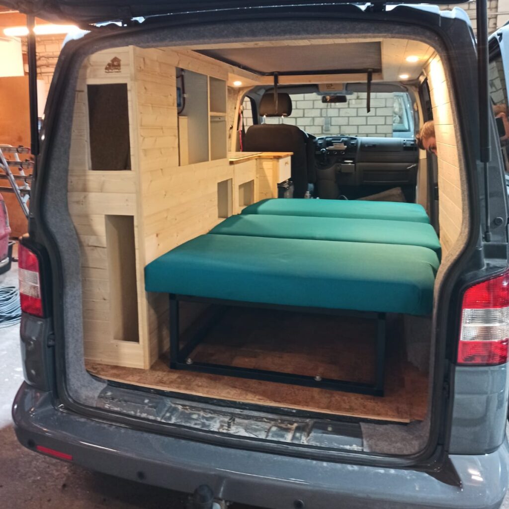 De bedbank is volledig geplaatst met petrol/groene kussens en uitgeklapt, gezien vanaf de achterkant door de achterklep die open staat