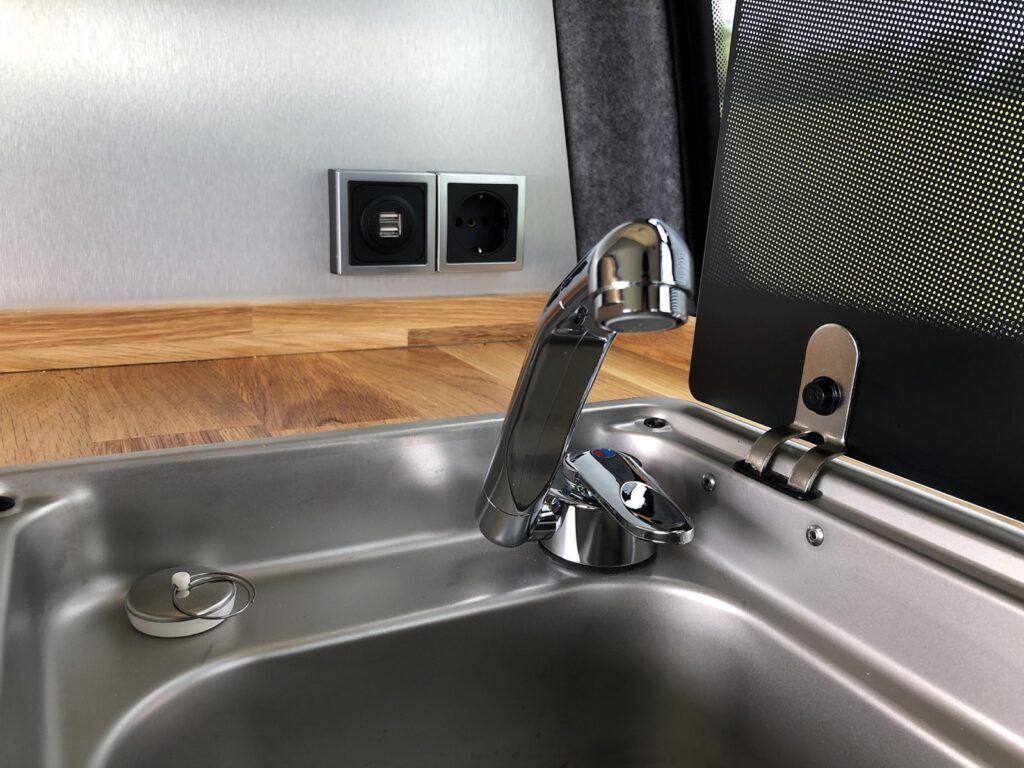 Close up van het verchroomde waterkraantje met daarachter op het grijze wandje een zwart stopcontact en twee usb-poorten